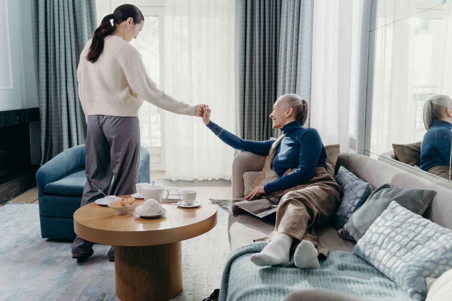 Betreuungskraft hilft einer älteren Dame beim Aufstehen im Wohnzimmer